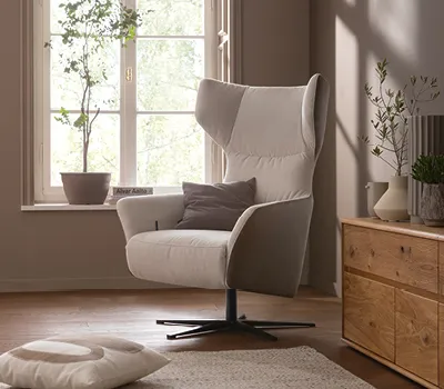 Gemütlicher Sessel in Beige und Grau mit Drehfuß, platziert vor einem Fenster in einem modernen Wohnzimmer mit Holzkommode und natürlicher Dekoration für entspannte Leseecke.